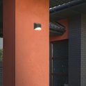 Donkergrijze buitenlamp gemonteerd op een oranje gestucte pilaar naast een donkergrijs bakstenen huis.