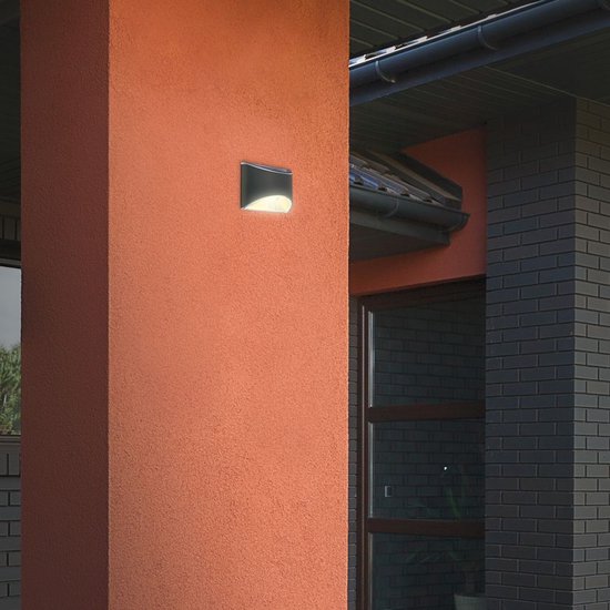 Donkergrijze buitenlamp gemonteerd op een oranje gestucte pilaar naast een donkergrijs bakstenen huis.