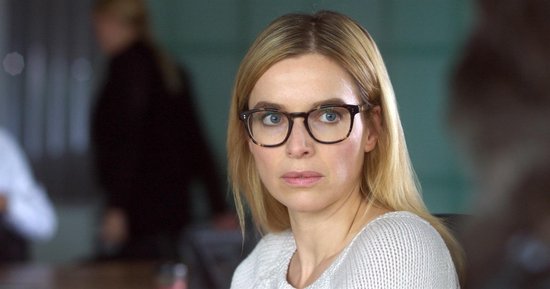 Een blonde vrouw met bril, gekleed in een wit gebreid vest, kijkt peinzend weg.