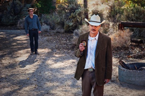 Man met cowboyhoed en bruin jasje, met een andere man op de achtergrond.