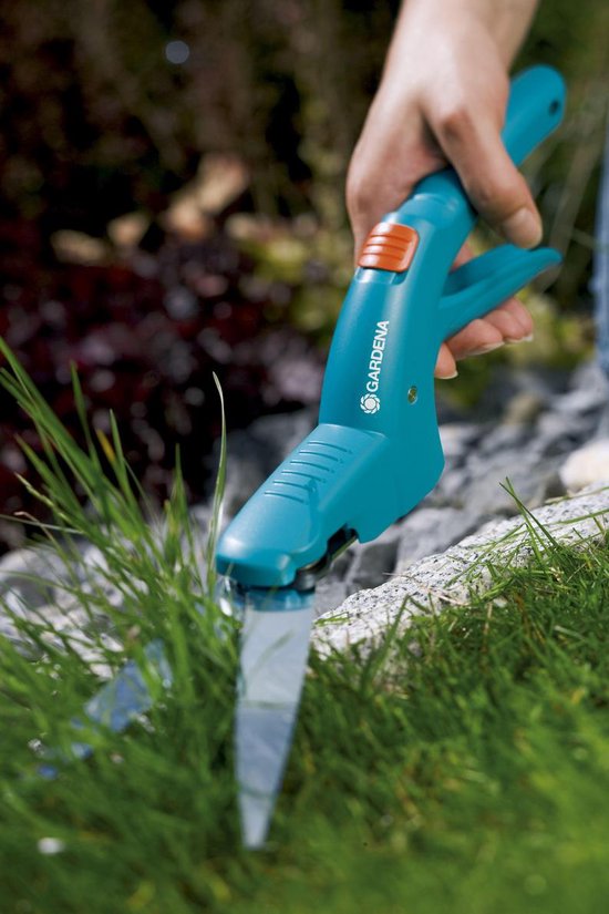 Een hand met een groen-blauwe Gardena grasschaar met oranje knop knipt gras.