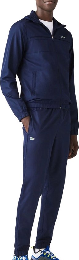 Een man draagt een marineblauw Lacoste trainingspak, bestaande uit een hoodie met rits en een trainingsbroek. Hij draagt witte sneakers.