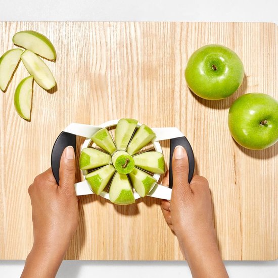 Des mains utilisent un coupe-pomme blanc avec des poignées noires pour découper une pomme sur une planche à découper en bois.