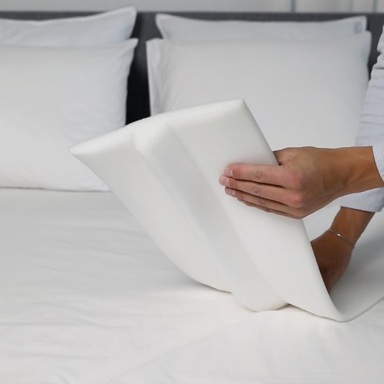 Hand houdt een witte, schuimrubberen matraswig met inkeping vast boven een bed.
