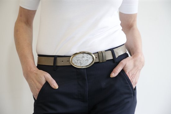 Bruine leren riem met ovale gesp met een beige-grijze mozaïekprint, gedragen door een model met een witte top en donkere broek.