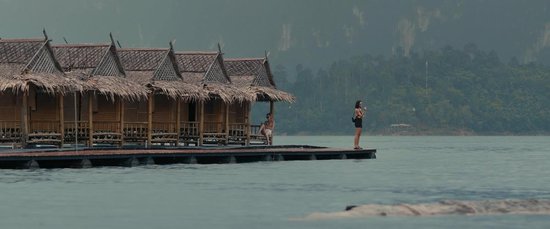 Een rij houten hutten met rieten daken op het water. Twee vrouwen staan op een steiger.