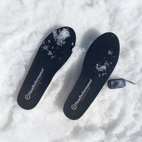 Twee zwarte HeatPerformance® inlegzolen met logo, bedekt met wat sneeuwvlokken, liggen in de sneeuw naast een kleine afstandsbediening.