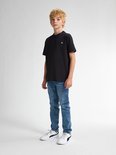 Jongen met blond haar staat in een zwarte t-shirt en blauwe jeans. Hij draagt witte sneakers met zwarte strepen.