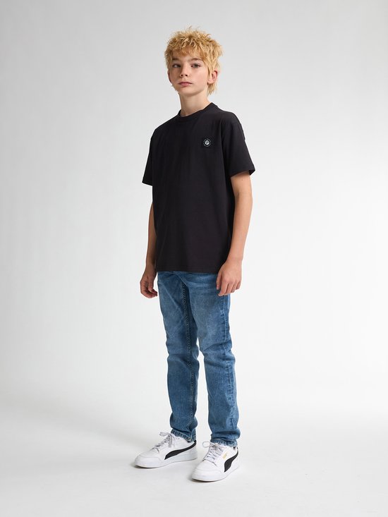 Jongen met blond haar staat in een zwarte t-shirt en blauwe jeans. Hij draagt witte sneakers met zwarte strepen.