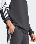 Man draagt een donkergrijze capuchontrui met witte mouwen en zwarte Adidas-strepen. Hij stopt zijn hand in zijn broekzak.