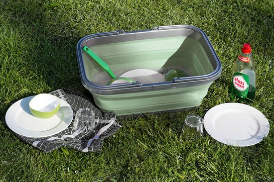 Groene opvouwbare wasteil met een groene borstel, gevuld met water en zeep, met borden en brillen ernaast op het gras.
