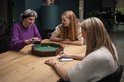 Drie vrouwen spelen een dobbelspel rond een achthoekige bak met groen vilt. Vijf dobbelstenen liggen in de bak.