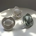 Drie Difrax Lovi fopspeen 18+ maanden in lichtgrijs, crème en saliegroen, op een witte tafel.