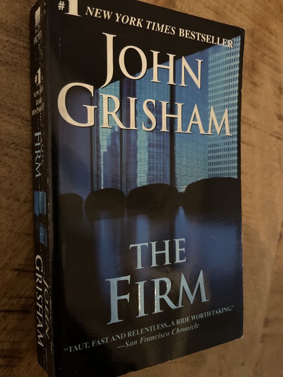 De zijkant van het boek 'The Firm' van John Grisham, met stadsgezicht op de achtergrond.