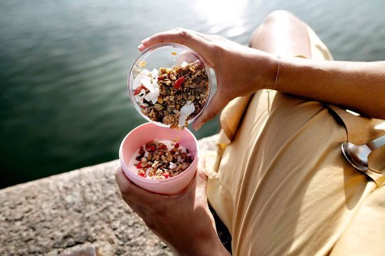 Vrouw gooit muesli van een transparante pot in een roze pot met yoghurt.