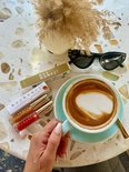 Een hand met nagellak houdt een gouden lipgloss flesje vast, naast een koffie met latte art, een zonnebril en een doosje.