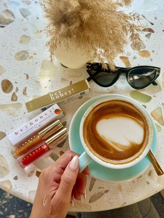 Een hand met nagellak houdt een gouden lipgloss flesje vast, naast een koffie met latte art, een zonnebril en een doosje.