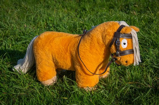 Oranje knuffelpony met bruin leren hoofdstel en witte manen, staand in het gras.