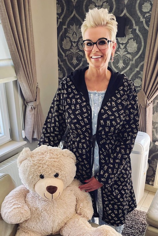 Une femme souriante aux cheveux blonds et portant des lunettes, se tient à côté d'un ours en peluche et porte un peignoir noir à imprimé ours en peluche.