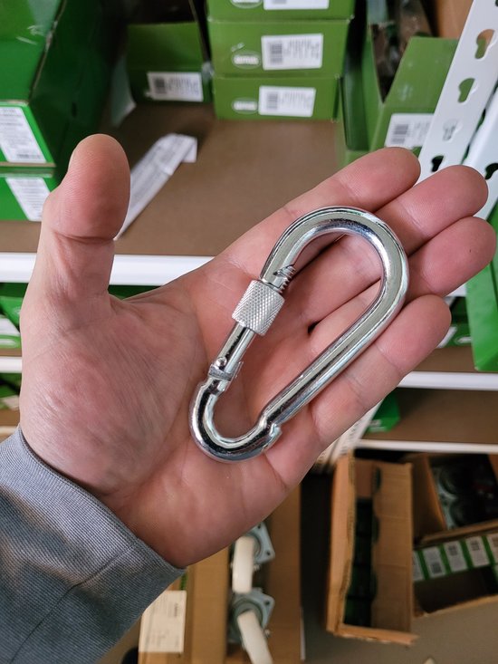 Een hand die een zilverkleurige karabijnhaak met schroefsluiting vasthoudt. Achtergrond met dozen.