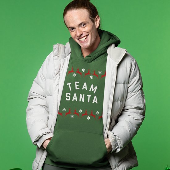 Model in groene kersthoodie met tekst 'TEAM SANTA' en rendier- en sneeuwvlokprint, gedragen onder een lichtgrijze jas.