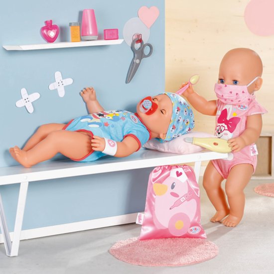 Een baby Born pop ligt op een bankje, een andere baby Born pop met masker geeft deze een thermometer.