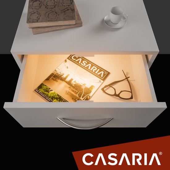 Open lade met verlichting, een tijdschrift met 'CASARIA' logo, brillen en boeken op een witte nachtkastje.