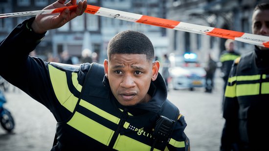 Politieagent met een geel hesje en blauwe uniform, kijkt gespannen naar links, afzetlint in beeld.