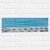 WallClassics - Muursticker - Rieten Parasols met Ligbedjes op het Strand - 60x20 cm Foto op Muursticker