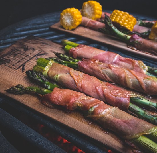 Groene asperges, omwikkeld met roze vleeswaren, op cederhouten planken boven een barbecue.