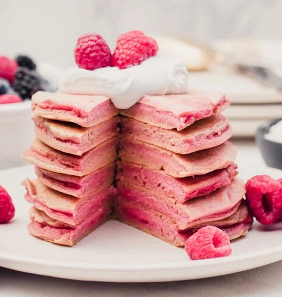 Stapel roze pannenkoeken met slagroom en frambozen.