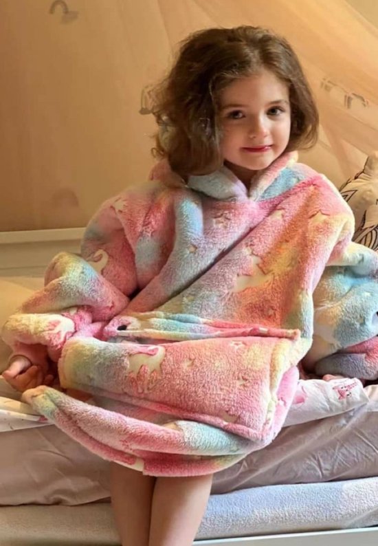 Een jong meisje met bruin haar zit op een bed, gekleed in een pluche hoodie deken met pastel regenboogkleuren, sterren en eenhoorns.