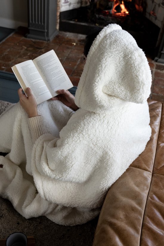 Een persoon in een witte teddy hoodie deken leest een boek, gezellig bij een haardvuur.