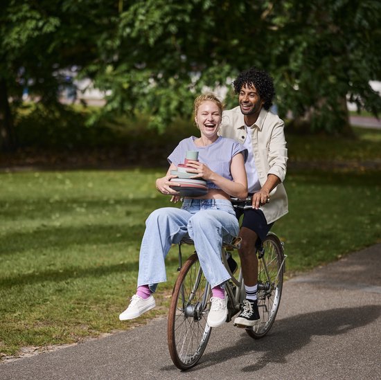 Een man en vrouw rijden op een fiets in een park, de vrouw draagt een stapel gekleurde mokken.