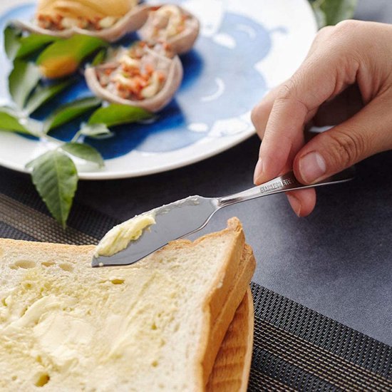 Hand die een zilverkleurig botermesje met 'STAINLESS STEEL' en 'House of Husk' inscriptie gebruikt om boter op toast te smeren.