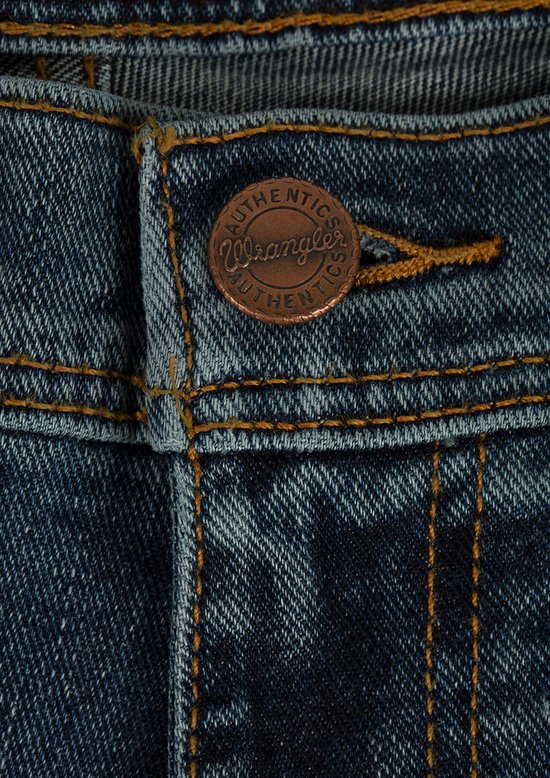 Detailopname van de koperkleurige Wrangler-knoop met opschrift Authentics op een blauwe spijkerbroek.