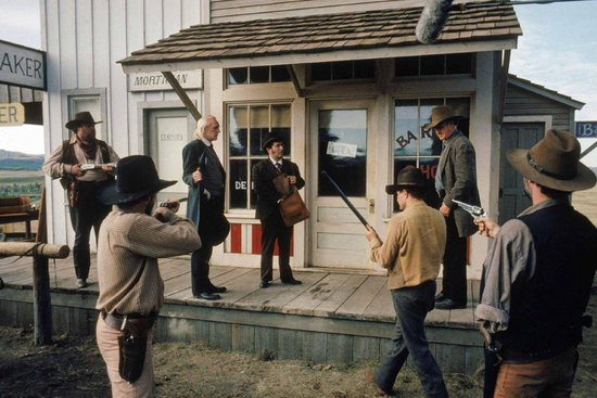 Een groep mannen met cowboyhoeden in een Western-dorp, sommigen met geweren, voor een gebouw met "BAKER" en "MORTIMER" erop.