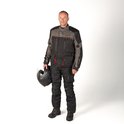 Man draagt een zwarte motorjas met donkergrijs en rode accenten en een motorbroek. Houdt een helm vast.