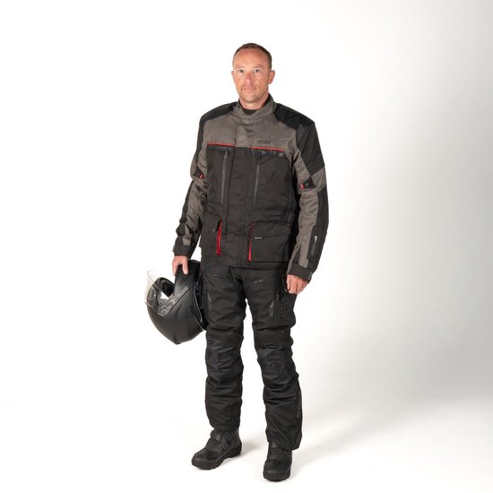 Man draagt een zwarte motorjas met donkergrijs en rode accenten en een motorbroek. Houdt een helm vast.