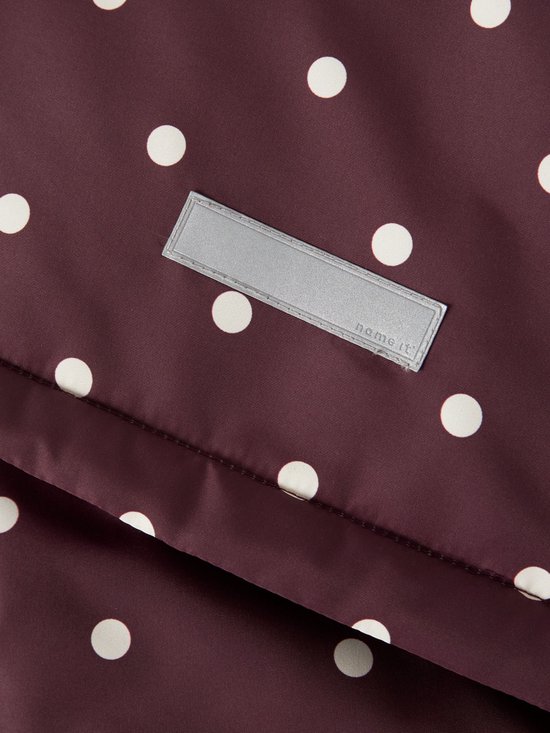 Detail van een label met 'name it' op een bordeauxrode jas met witte stippen.