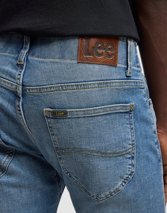 Close-up van de achterkant van een spijkerbroek met een bruin leren Lee-patch op de tailleband en een klein Lee-logo op de rechterzak.