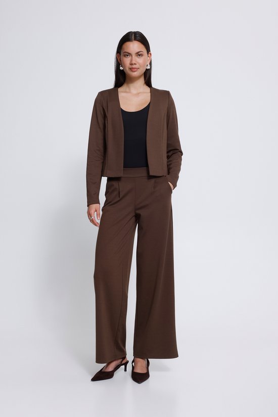 Model in een bruine wijde broek en bijpassend cropped jasje, met een zwarte top eronder. Bruine puntige schoenen.