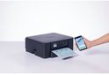 Een hand houdt een smartphone met een geprinte reiskaart omhoog, naast een zwarte Brother printer.