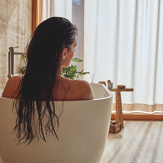 Vrouw met lang, donker, nat haar ontspant in een wit bad met een lichte achtergrond.