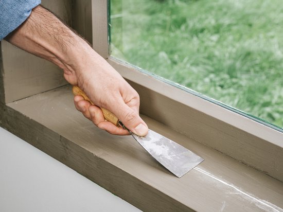 Hand gebruikt een plamuurmes om witte vulmiddel uit te strijken op een houten vensterbank.