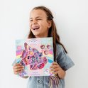 Lachend meisje met open mond houdt een knutseldoos met eenhoorntje omhoog. Doos bevat veel gekleurde kralen en tassels.