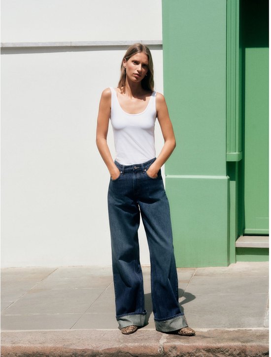 Model in een donkerblauwe boyfriend jeans met omgeslagen pijpen en een witte tanktop.