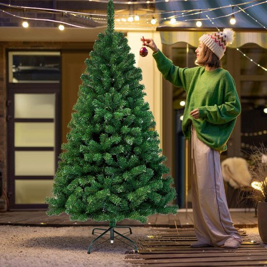 Vrouw met gebreide muts en groene trui hangt een rode kerstbal op in een groene kunstkerstboom.