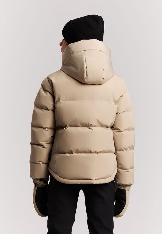 Een jong meisje met blonde haren staat van achteren in een beige winterjas, zwarte muts en zwarte skibroek.