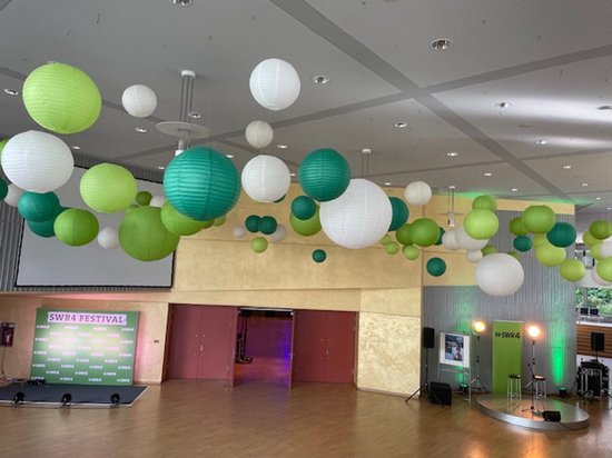 Veel ronde papieren lampionnen in wit, lichtgroen en donkergroen hangen aan het plafond. Op de achtergrond een podium met een scherm.